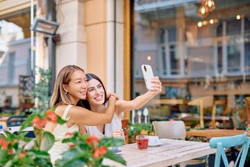 Two female friends are taking a selfie with a smartphone while sitting at a table in an outdoor cafe, enjoying their time together