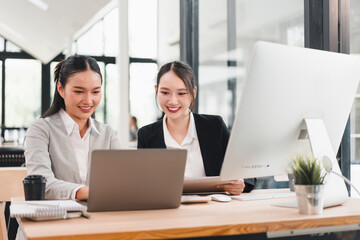 Two Asian businesswomen, smiling and working together at modern office desk, using laptop and digital tablet, with desktop computer and coffee cup, teamwork and collaboration concept