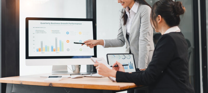 Two businesswomen formal attire discussing quarterly business growth performance modern office, with one pointing computer screen displaying colorful bar and line charts, teamwork and analysis