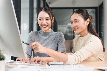 Obraz premium Two young Asian women working together at desk, smiling and looking at computer screen in modern office, collaborating on project with positive energy