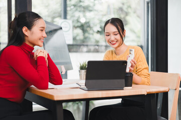 Two young Asian women, one in red sweater and other in yellow sweater, sit at wooden table in modern office, smiling and discussing work with digital tablet and documents