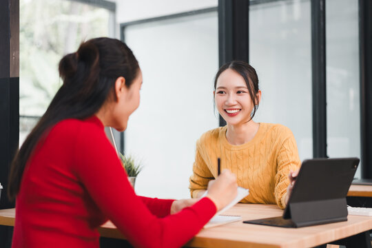Two Asian women, one in yellow sweater and one in red sweater, sit at wooden table in modern office, smiling and talking, with digital tablet and notebook present