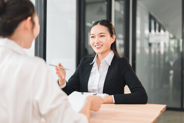 Two businesswomen having friendly conversation in modern office setting, one smiling and holding...