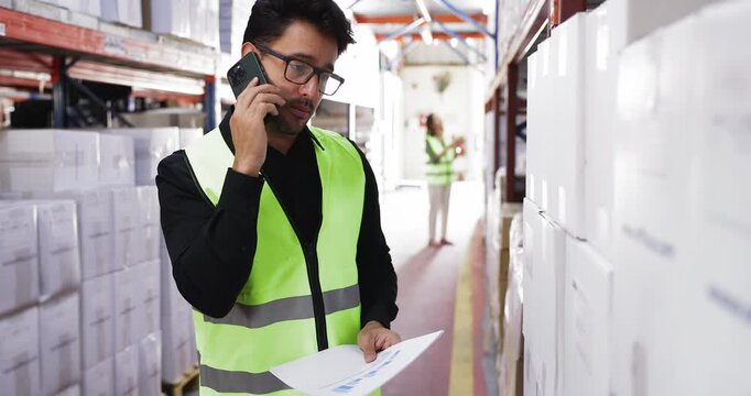 Hispanic male, warehouse or logistics coordinator worker dressed in safety vest discussing inventory data, shipping or supply chain operations on smartphone. Warehouse, distribution center workflow