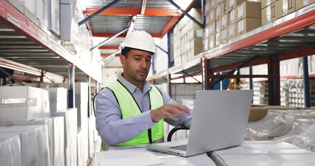 Young 35s focused Hispanic man working in well-organized warehouse, wear protective helmet and vest reviewing inventory documents while working on laptop. Logistics, inventory management, supply chain - Powered by Adobe