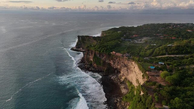 Aerial drone footage gliding over Pura Luhur Uluwatu dramatic cliffs, lush green hills, and a serene beach bathed in the golden light of sunset. Bali, Indonesia