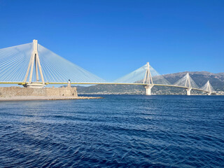 This photo shows the Rion-Antirion Bridge in Greece, stretching across deep blue waters with stunning mountains in the background. A clear sky and bright light emphasize its modern, elegant design.