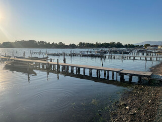 This photo features a rustic wooden dock leading to a small marina with fishing boats. Calm, clear...