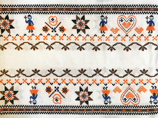This vibrant traditional textile features cross-stitched patterns with hearts, stars, flowers, and folk dancers in blue and orange tones. It reflects rich cultural heritage and handcrafted artistry.