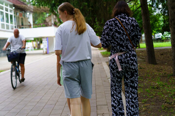 Two women walk along a pathway while a man rides a bicycle in a green park during a sunny day in...