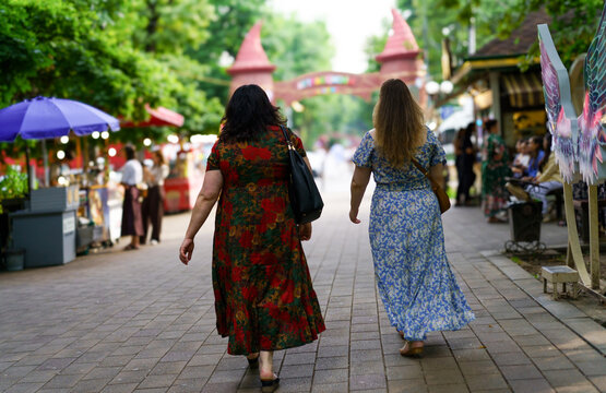 Two women stroll through a vibrant market in a lively outdoor setting during a sunny afternoon - Powered by Adobe