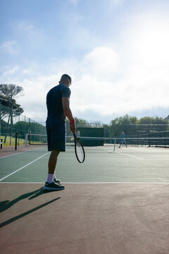 Male tennis player standing near net holding racket facing opponent on fenced park court
