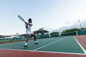 Diverse male athletes in white sportswear swinging rackets at tennis ball on outdoor court with net