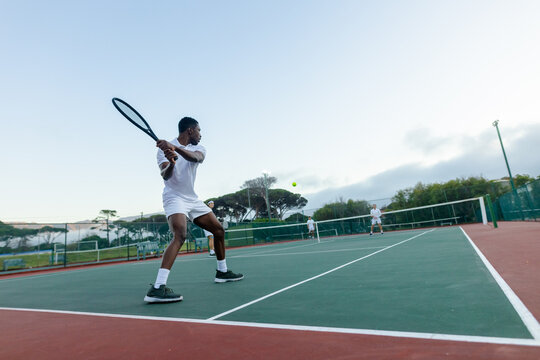 Diverse male athletes in white sportswear swinging rackets at tennis ball on outdoor court with net