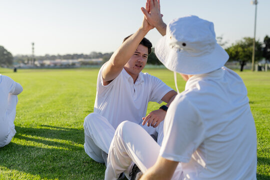 Two male cricket teammates sitting on field high-fiving each other in cricket uniforms, bucket hat - Powered by Adobe