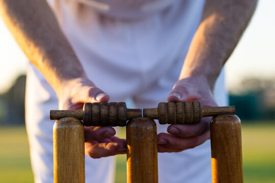 Cricket player placing wooden bail on worn stumps on grassy pitch under warm sunlight