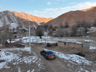 A black car drives along a snowy dirt road in a rustic mountain setting, with a small adobe...