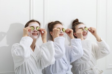 Happy women with facial masks and cucumber slices in bathrobes after spa procedures near white wall