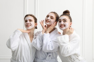 Happy women with facial masks in bathrobes posing after spa procedures near white wall