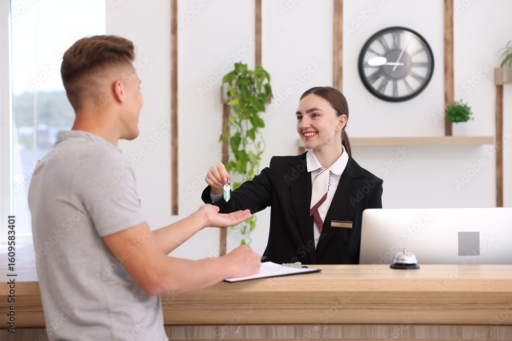 Poster smiling receptionist in uniform giving guest room key to client at reception desk in hotel