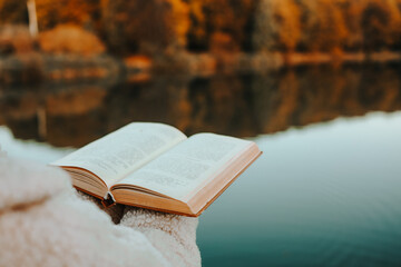 Person reading a book near calm lake in autumn, with golden trees reflecting in water, evoking...