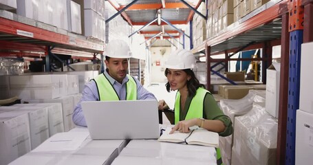 Two warehouse workers wearing protective uniforms working together using laptop, checking and updating stock information, reviewing new orders, engaged in inventory management, verify goods quantities - Powered by Adobe