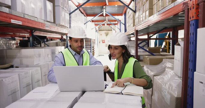 Two warehouse workers wearing protective uniforms working together using laptop, checking and updating stock information, reviewing new orders, engaged in inventory management, verify goods quantities