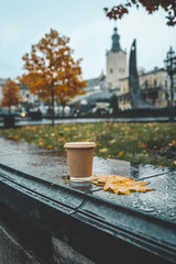 Takeaway coffee cup on a wet stone bench with autumn leaves in the foreground. City park with blurred yellow trees and historical architecture in the background