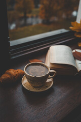 Cozy autumn scene with a ceramic cup of coffee, a croissant, and an open book on a windowsill, with golden leaves and blurred city buildings in the background