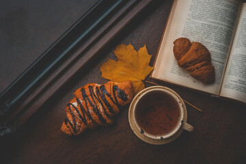 Flat lay of a cozy autumn breakfast with a cup of tea, croissants, open book, and yellow maple leaf...
