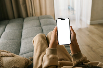 Woman in cozy beige outfit lying on sofa and holding smartphone with blank white screen. Relaxing at home in soft natural light through curtains