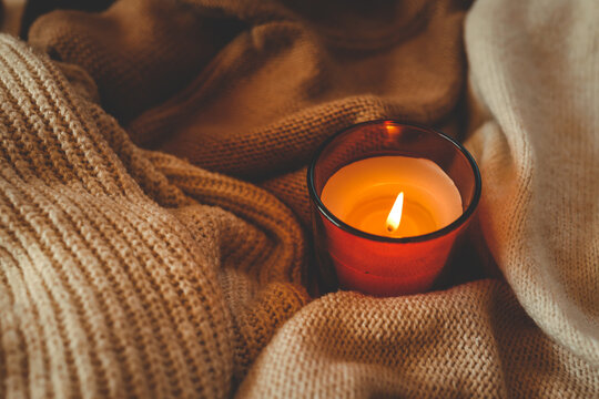 Top view of a burning candle on cozy knitted blankets in warm beige and brown tones. Soft textures and warm light create a relaxing autumn or winter atmosphere
