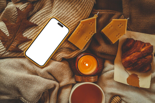 Cozy autumn flat lay with smartphone, beeswax house candles, cup of tea, burning candle, croissant and dry leaves on soft beige knitted blankets - Powered by Adobe