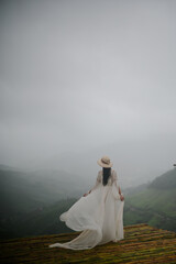 A woman in a flowing white dress and hat stands on a wooden platform, overlooking misty, green...