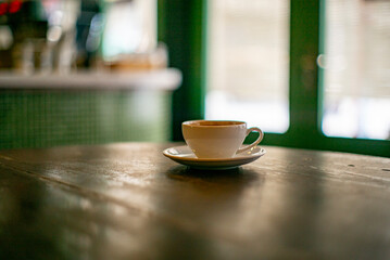 A close-up of a white coffee cup and saucer rests on a dark, textured wooden table, captured with a shallow depth of field. The softly blurred background reveals green tiled walls and window
