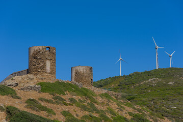 Ancient stone ruins stand on a rugged green hillside alongside modern white wind turbines under a...