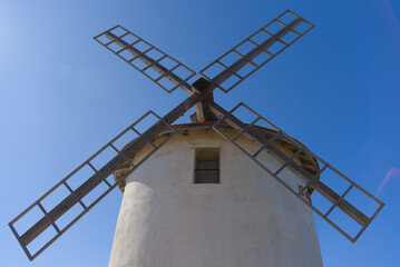 A historic white windmill with a red-tiled conical roof and wooden sails stands atop a green, bushy...
