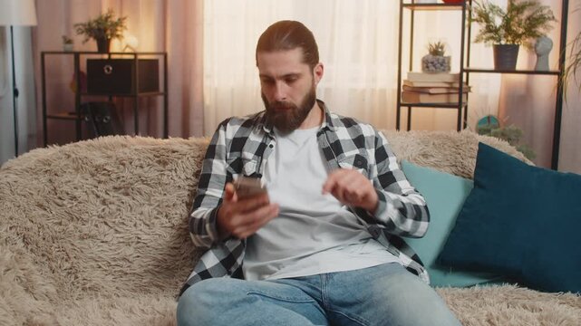 Young man sitting at home, receiving crucial smartphone call quarrel and placing device on knee with visible tension. Caucasian guy gestures irritably, pushing a firm argument through raised voice.