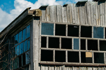 An old wooden abandoned house with broken windows stands in a field among overgrown grass on a summer day against a clear and clear blue sky with clouds. the texture of an old house