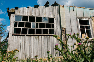 An old wooden abandoned house with broken windows stands in a field among overgrown grass on a summer day against a clear and clear blue sky with clouds. the texture of an old house