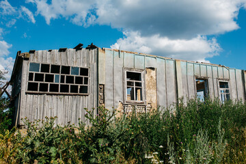 An old wooden abandoned house with broken windows stands in a field among overgrown grass on a summer day against a clear and clear blue sky with clouds. the texture of an old house