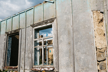 An old wooden abandoned house with broken windows stands in a field among overgrown grass on a summer day against a clear and clear blue sky with clouds. the texture of an old house