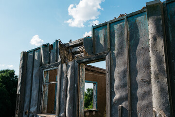 An old wooden abandoned house with broken windows stands in a field among overgrown grass on a summer day against a clear and clear blue sky with clouds. the texture of an old house