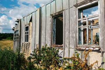 An old wooden abandoned house with broken windows stands in a field among overgrown grass on a summer day against a clear and clear blue sky with clouds. the texture of an old house