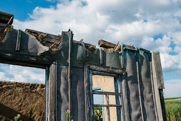 An old wooden abandoned house with broken windows stands in a field among overgrown grass on a summer day against a clear and clear blue sky with clouds. the texture of an old house