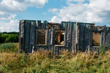 An old wooden abandoned house with broken windows stands in a field among overgrown grass on a summer day against a clear and clear blue sky with clouds. the texture of an old house