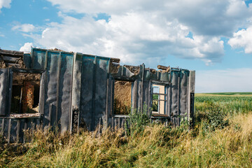 An old wooden abandoned house with broken windows stands in a field among overgrown grass on a summer day against a clear and clear blue sky with clouds. the texture of an old house