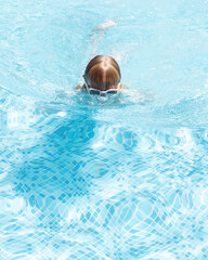 Girl rising from blue swimming pool water, wet hair shining in sun. Vertical action shot in outdoor swimming pool. Concept of swim, water sports, training, summer activities, children's sport