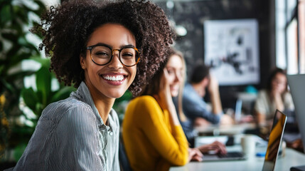 A smiling young woman with curly hair and glasses in a modern office setting, surrounded by colleagues working on laptops.