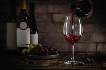 A dark, elegant still life of red wine being poured into a glass with wine bottles and fresh grapes...